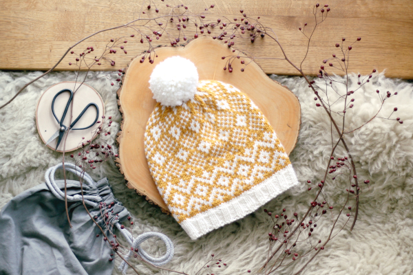 Czapka Goldenrod Hat, biało żółta z pomponem, ułożona na drewnianej podkładce, otoczona suszonymi gałązkami.